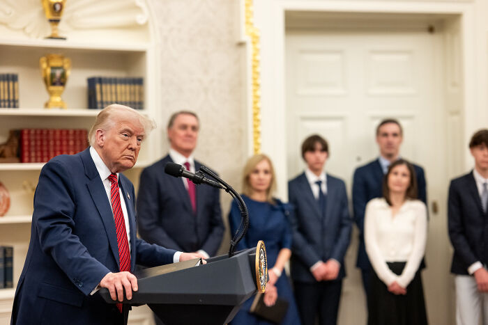 Donald Trump at a podium speaking in a formal room with people standing behind him during a D.C. event.