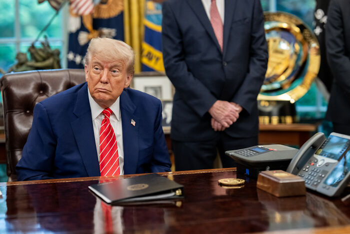 Donald Trump sitting at desk in the Oval Office during a formal meeting with people standing behind him