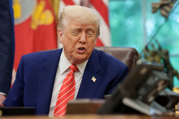 Donald Trump seated in office wearing a navy suit and red striped tie discussing Qatar Royals flying palace for Air Force One deal.