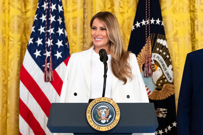 Melania Trump smiling at a podium with US flags behind, relating to Trump and Melania separated marriage norms.