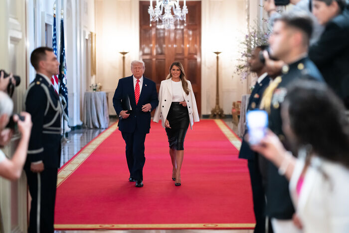 Donald Trump and Melania walking down a red carpet in a formal setting with military personnel and photographers around.