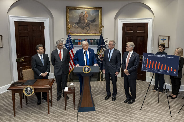 Donald Trump speaking at a podium with officials and a chart, discussing fat shot and vaccine costs abroad.