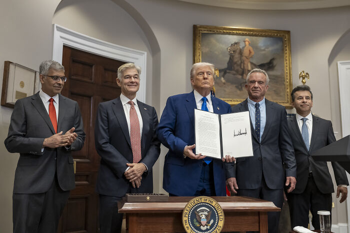 Donald Trump in a suit holding a signed document, standing with four men in suits in a formal room with presidential seal and painting.
