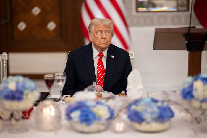 Donald Trump sitting at a formal dining table with blue floral centerpieces and an American flag in the background.