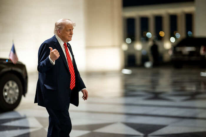 Donald Trump walking outside near a vehicle at night, with blurred lights in the background.