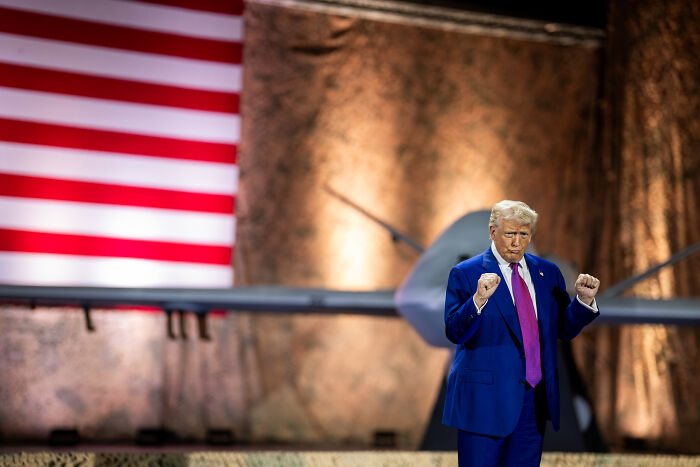 Donald Trump in a blue suit, addressing a crowd with an American flag and aircraft in the background during a public event.