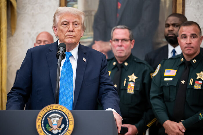 Donald Trump speaking at a podium with law enforcement officers behind him, addressing tariffs and legal challenges.
