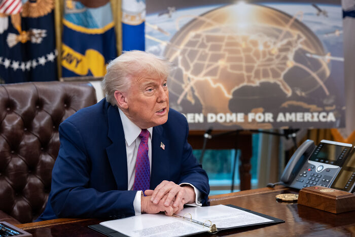 Former President Trump sitting at a desk discussing a bold defense deal with Canada if it joins as the 51st state.
