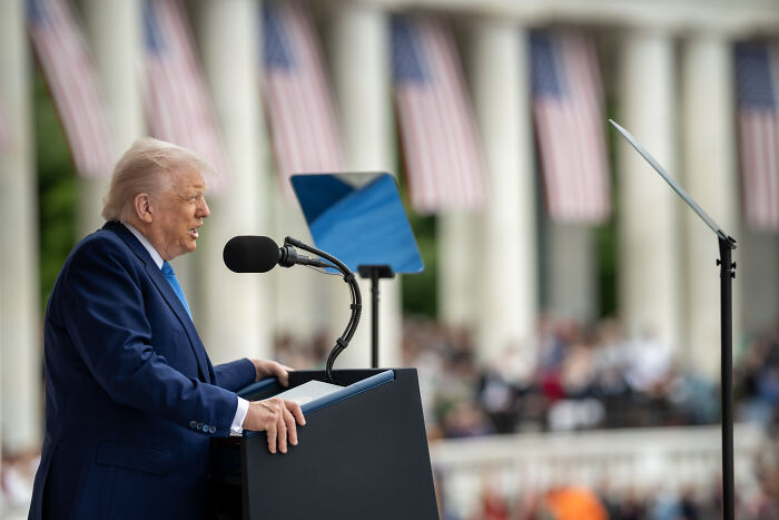 Man in suit speaking at podium with microphone and teleprompter, with American flags in the background. Student visa freeze.