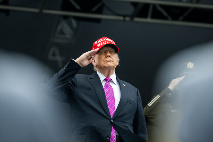Donald Trump wearing a red Make America Great Again hat, saluting during a public event.