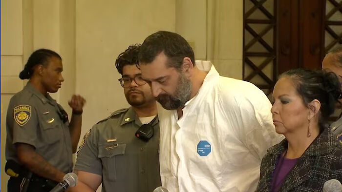 Man allegedly beat 12-year-old son to death with baseball bat, shown in custody with law enforcement officers present.