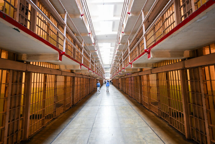 Interior view of Alcatraz prison cells lining a long corridor with a few visitors walking inside the notorious facility.