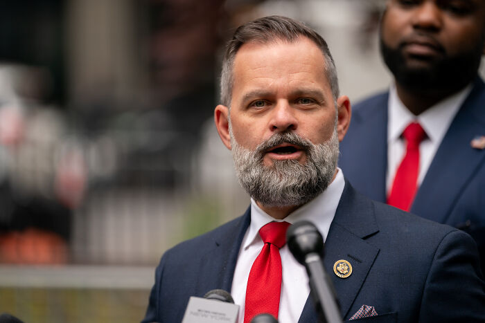 Man with beard and red tie speaking at microphone, linked to Republican accused of lying about military service and Bronze Star.