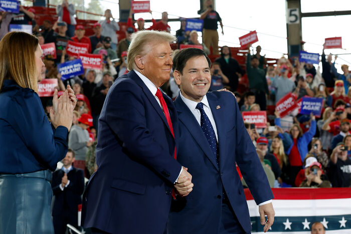Donald Trump shaking hands with a man at a rally, with supporters holding signs in the background about renaming a Gulf.