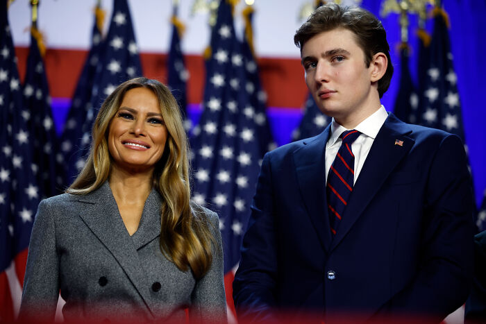 Melania Trump and Barron Trump standing in front of American flags at a formal event related to college rumors.