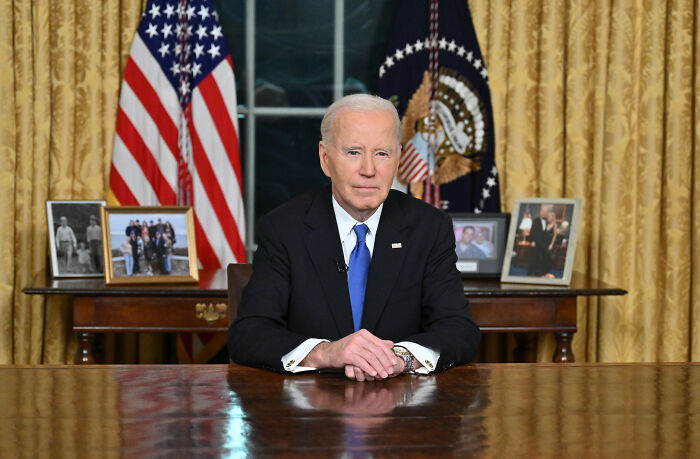 Joe Biden sitting at desk in Oval Office, with US flags and family photos behind, amid cancer diagnosis conspiracy theories.