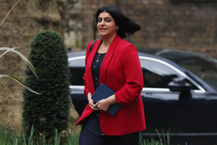 Woman wearing a red blazer walking outdoors next to trimmed bushes and a black car, related to child exploiters crackdown news.