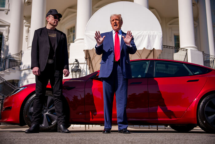 Elon Musk wearing sunglasses and black outfit standing next to a red car with another man in a blue suit speaking