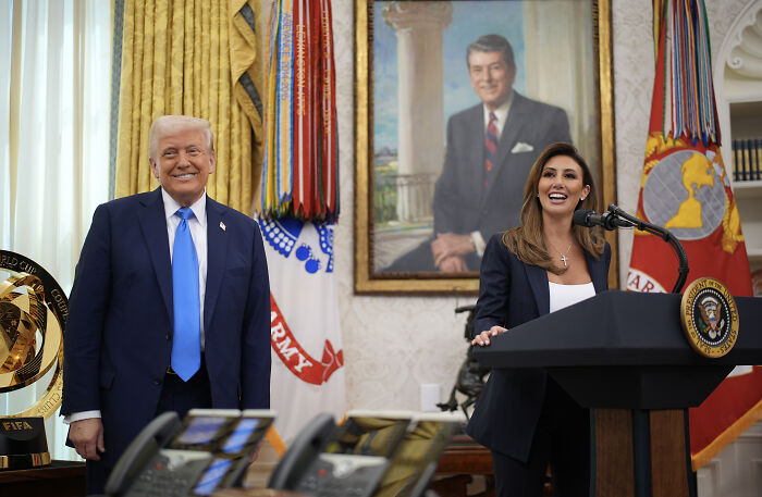 Former President Donald Trump standing beside a woman speaking at a presidential podium in the Oval Office.