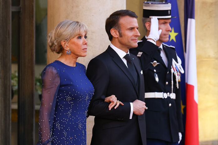 Brigitte Macron and Emmanuel Macron standing together at a formal event with French and European Union flags behind them.