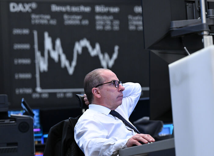 Man in glasses and white shirt monitoring financial data on multiple screens with stock market graph in background.