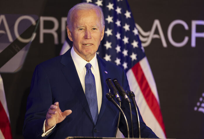 Joe Biden speaking at a podium with American flag in background amid cancer diagnosis cover-up claims.