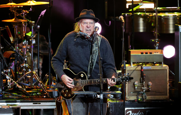 Neil Young playing guitar on stage wearing a hat, focusing on musicians amid political controversy.