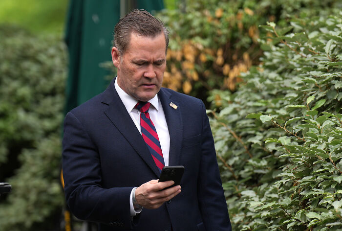 Mike Waltz in a dark suit and striped tie looking at his phone outdoors amid green foliage.