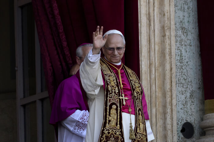 New Pope Leo XIV in ceremonial robes waving, linked to accusations of overlooking child abuse claims in Chicago parish.