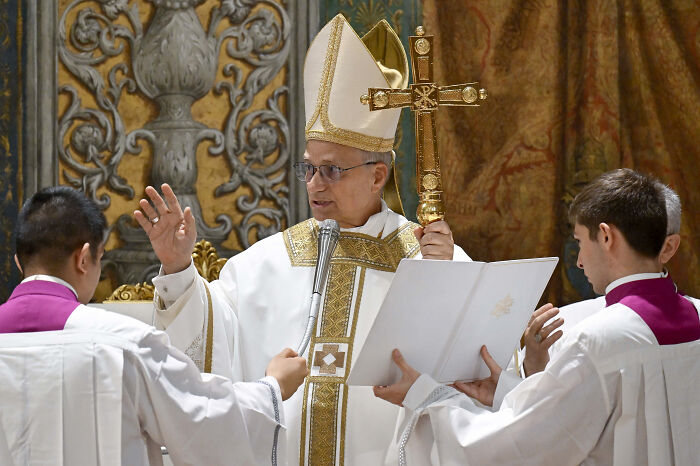 Pope Leo XIV holding a crucifix and speaking during a religious ceremony, flanked by two altar servers.