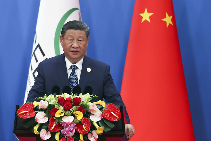 Xi Jinping speaking at a podium with floral arrangement, Chinese flag, and global trade discussion context.