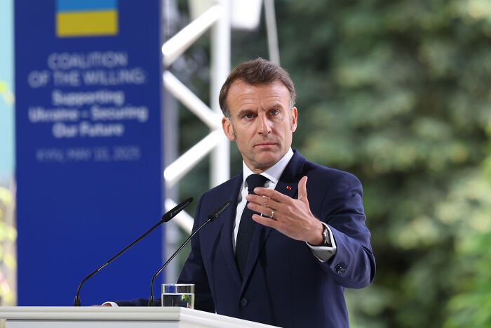 Emmanuel Macron speaking at a podium outdoors, wearing a dark suit and gesturing with his hand during a speech.