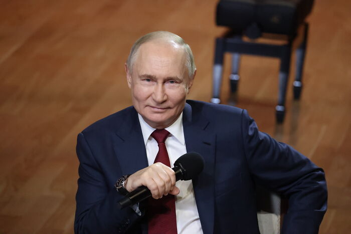 Vladimir Putin in a dark suit and red tie holding a microphone, speaking during a formal event indoors.