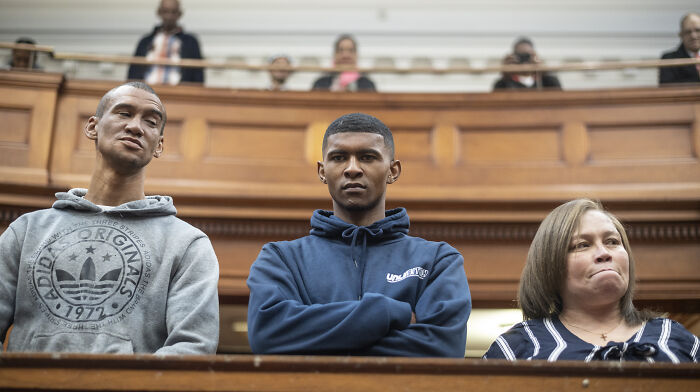 Three people in a courtroom setting, with a woman, two men, and wooden paneling in the background.