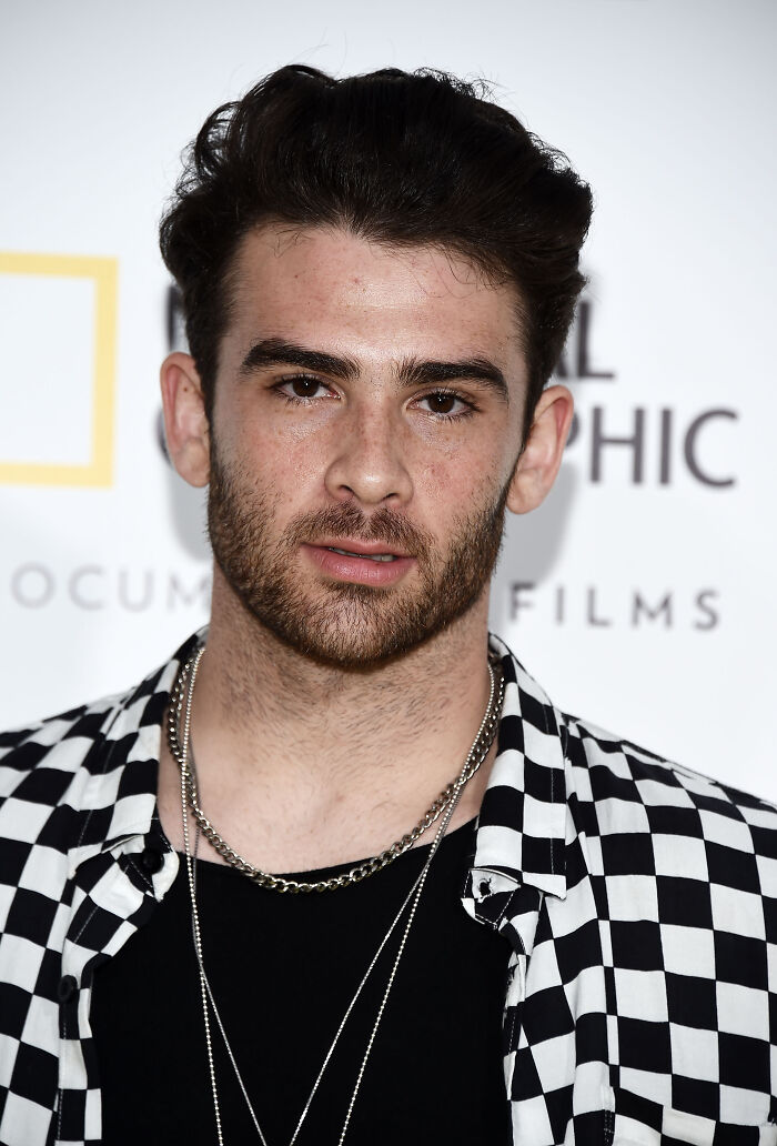 Young man with dark hair and beard wearing checkered shirt and chains in front of a white background with logos popular left-wing Twitch streamer Hasan Piker