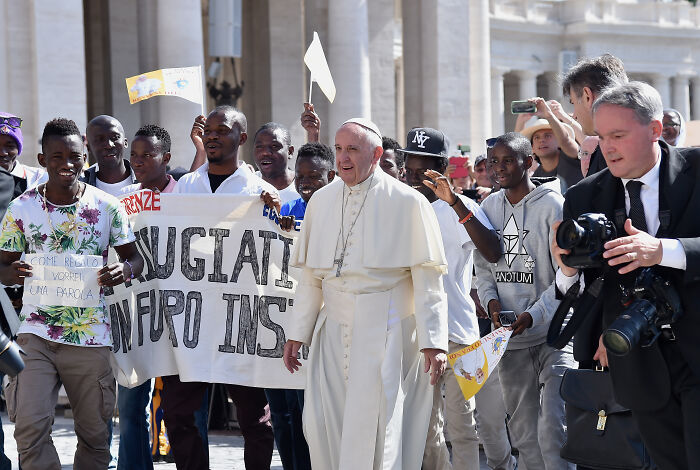 Pope Leo XIV walking with supporters holding signs on immigration and refugees in a public square.