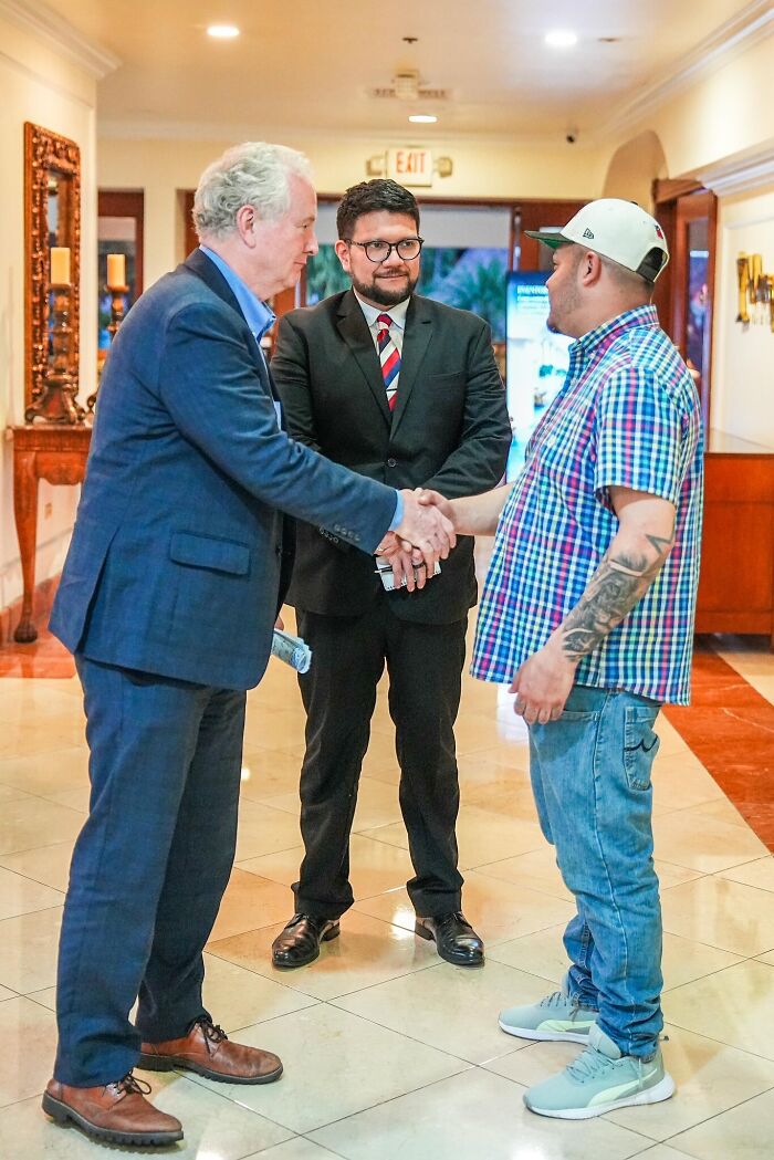 Two men shaking hands while a third man in a suit watches, discussing deportees in a formal indoor setting.