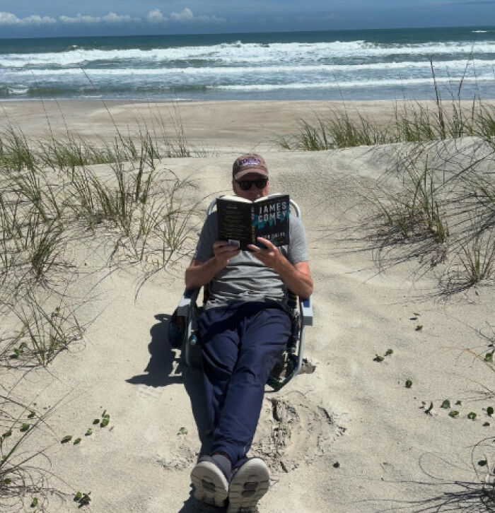 Man wearing sunglasses and cap relaxing on beach chair reading a James Comey book near ocean waves.