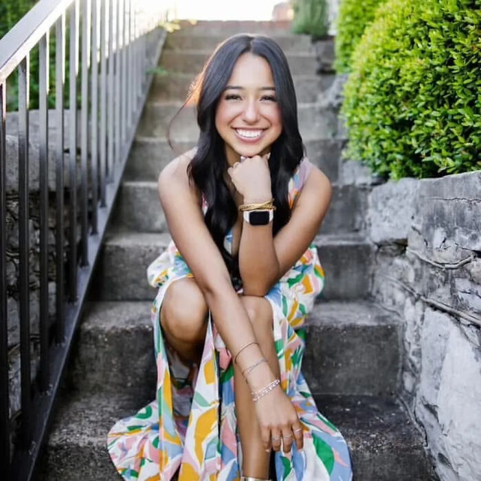 Young college student sitting on outdoor steps, smiling, wearing colorful dress, related to deportation case after illegal turn at red light.