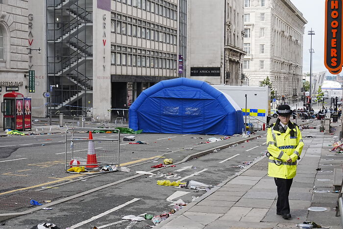 Police officer walking near a blue police tent on a littered street after vandalism and Liverpool FC parade incident.