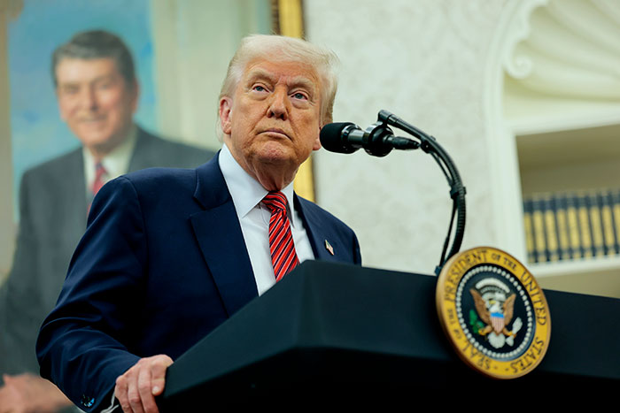 Donald Trump at a presidential podium, speaking in a formal room with a historic portrait in the background.