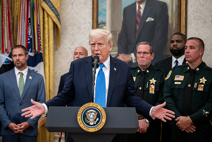 Donald Trump speaking at a podium with security officials nearby, biographer explores bitterness toward Harvard.