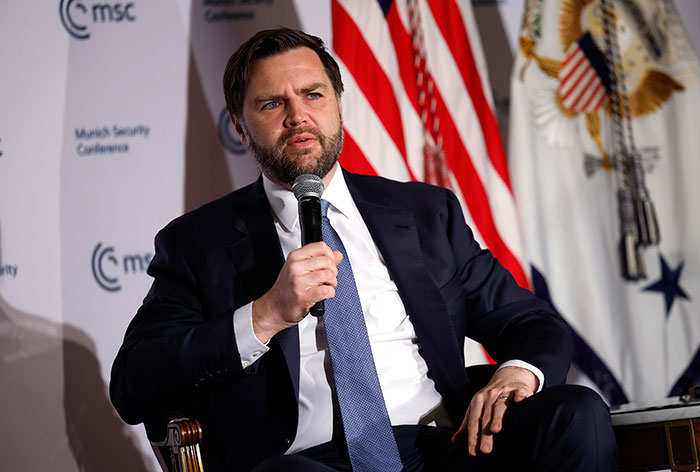 JD Vance speaking at a conference, holding a microphone with American flag and presidential seal in the background