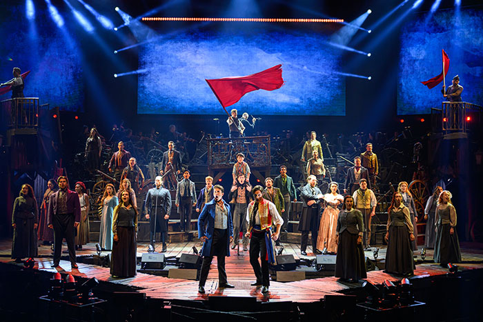 Les Mis&eacute;rables cast performing on stage holding red flags during a dramatic theater production.
