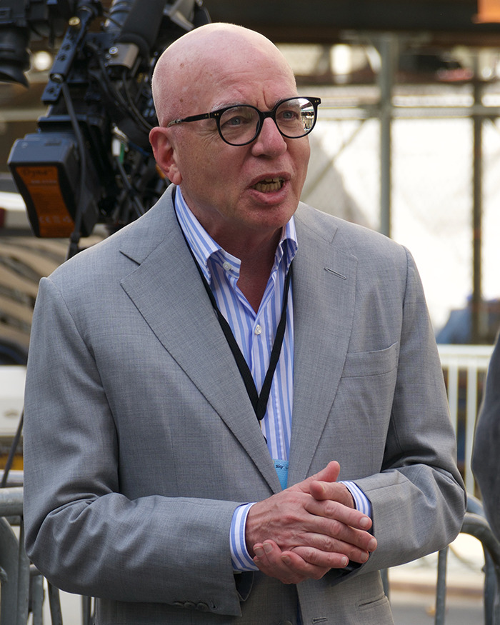Biographer speaking outdoors, wearing glasses and a gray suit, discussing the real reason Donald Trump feels bitter toward Harvard.