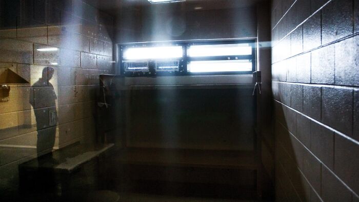 Dimly lit prison cell with barred window and shadow of a person, reflecting private prison and migrant crackdowns.