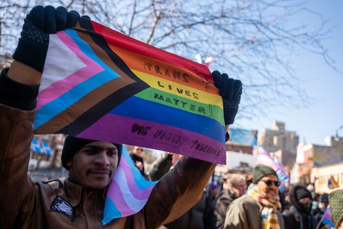 Person holding a trans pride flag with "Trans Lives Matter" at a protest, symbolizing trans troops&rsquo; rights amid legal battles.