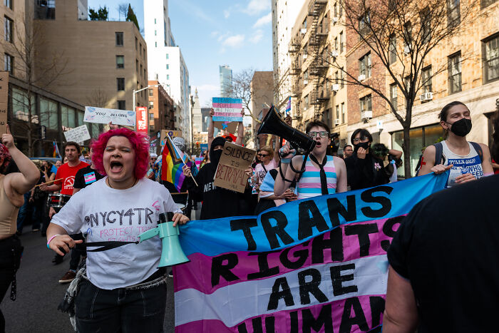 Protesters holding trans rights banner and megaphones, advocating for trans troops amid court legal battle.