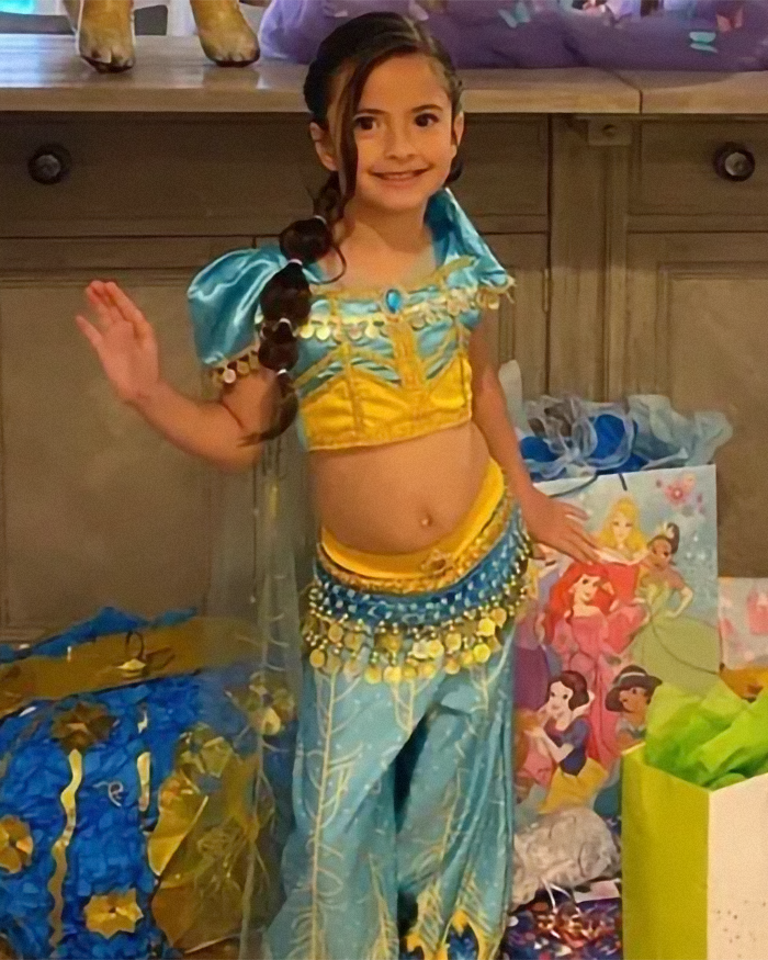 Young girl in a colorful princess costume smiling and waving, surrounded by gift bags and birthday decorations.
