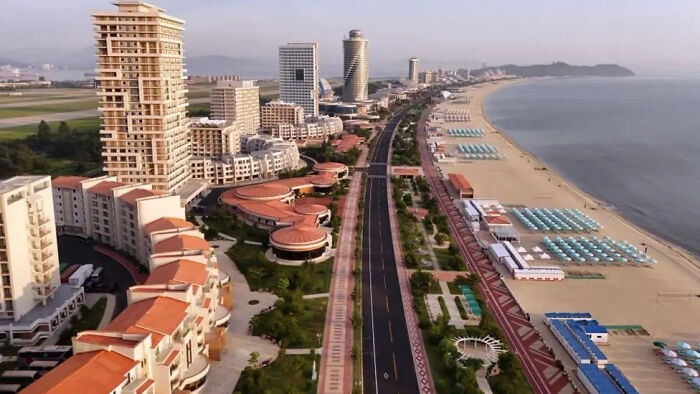 Aerial view of North Korea beach resort with buildings, empty road, and rows of blue beach umbrellas by the ocean.